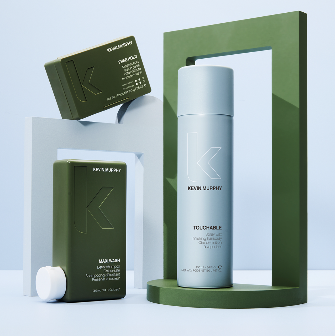 Product shot of Kevin Murphy hair products including shampoo, styling cream, and hairspray on a minimalist geometric stage against a blue background.
