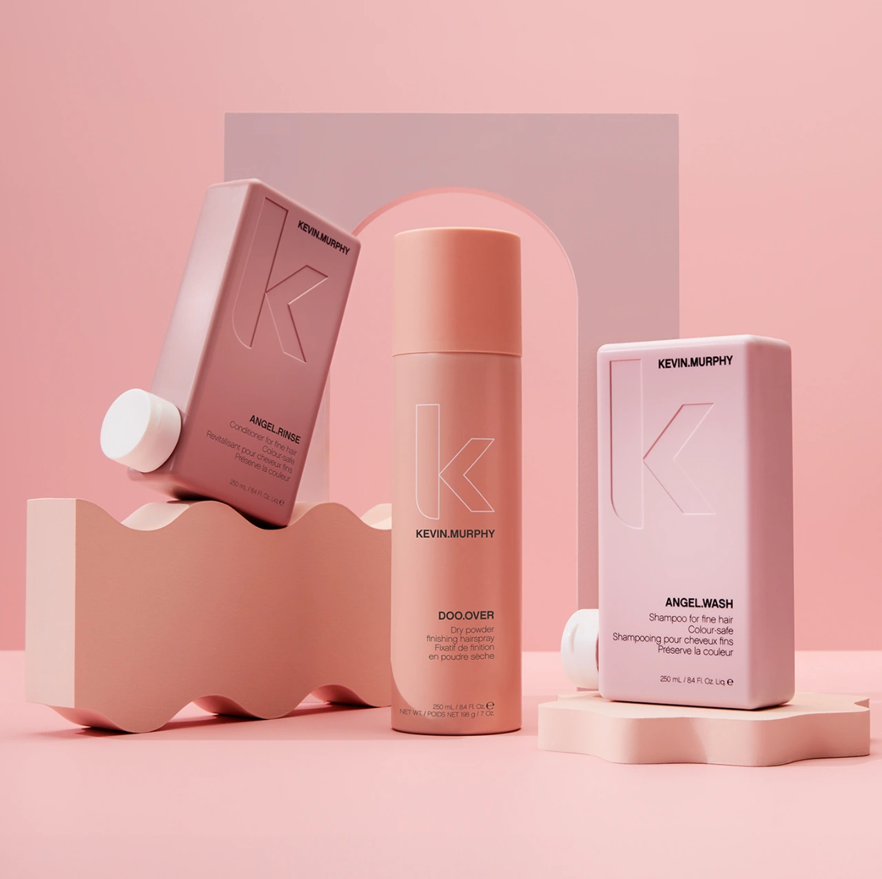 Three Kevin Murphy hair product bottles arranged on pink stylized pedestals against a soft pink arched studio backdrop.