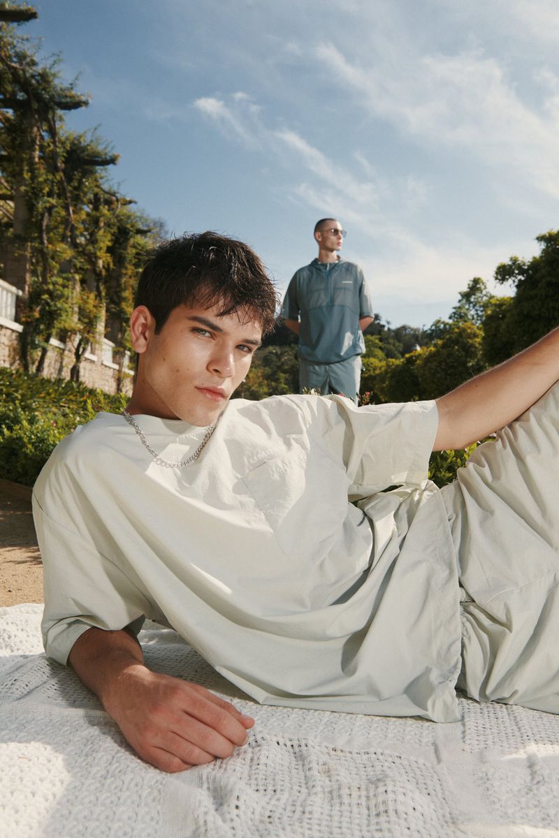Two adult males pose outdoors on a blanket. One man lies on a white crocheted blanket, wearing a light-grey T-shirt and shorts.