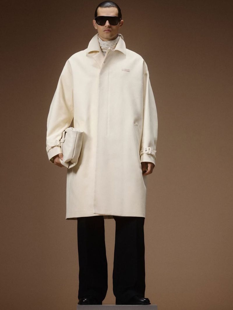 Male model in an oversized off-white coat, dark shield sunglasses, and wide-leg black trousers on a brown backdrop.