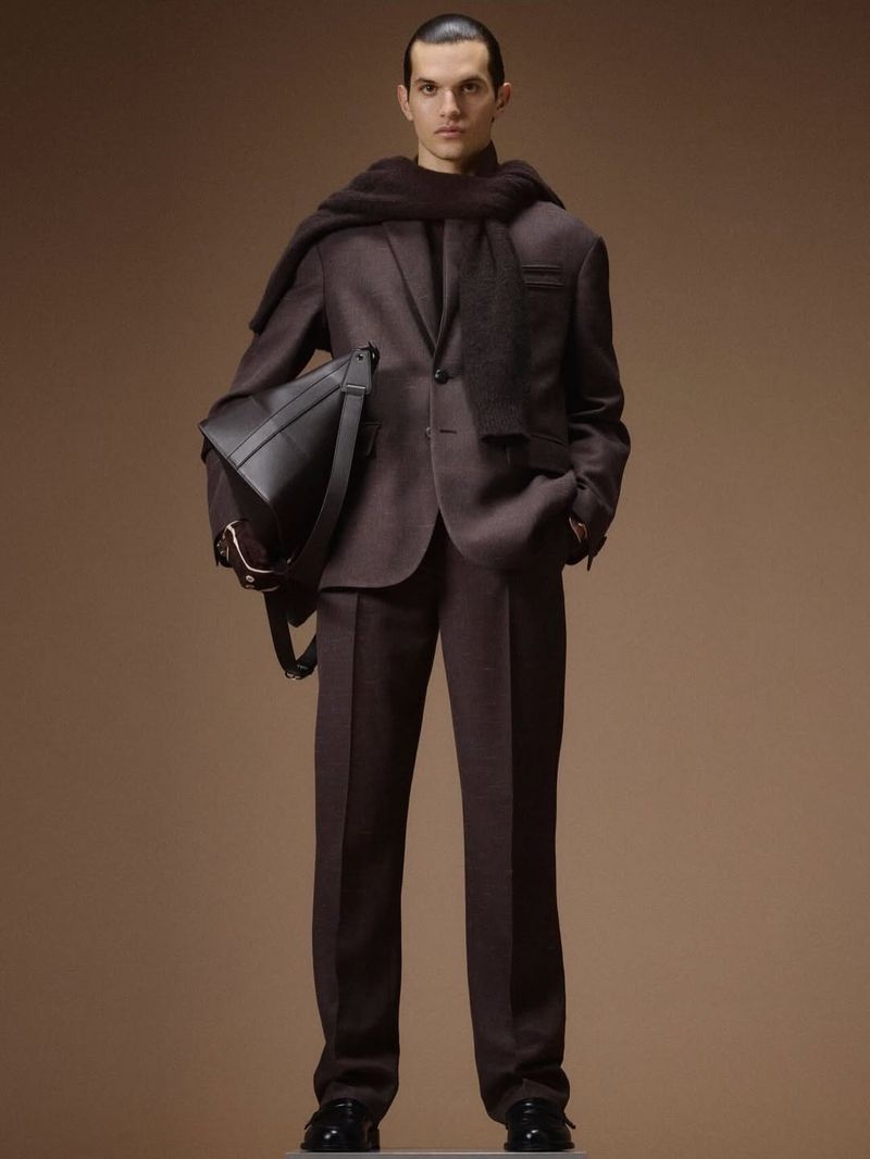 Male model wearing a dark brown tailored suit, draped wool scarf, and black leather shoulder bag against a warm studio backdrop.