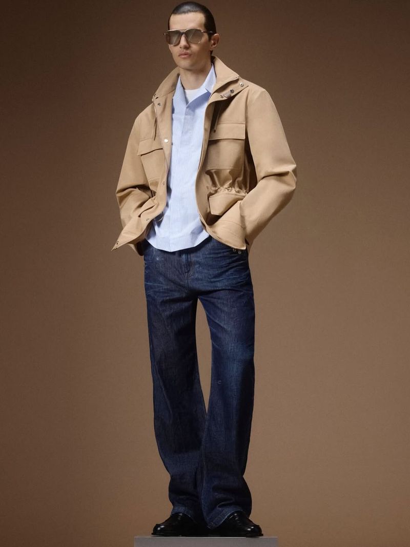 Model wearing a tan utility jacket, light blue shirt, and dark wide-leg denim jeans against a warm brown studio backdrop.