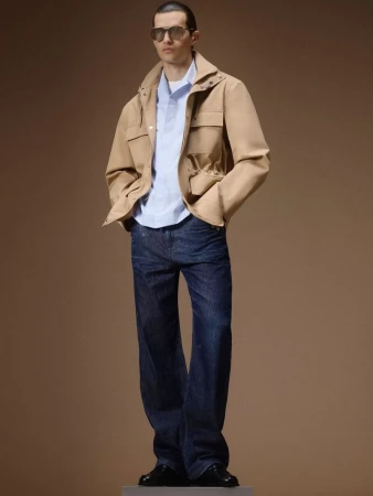 Model wearing a tan utility jacket, light blue shirt, and dark wide-leg denim jeans against a warm brown studio backdrop.