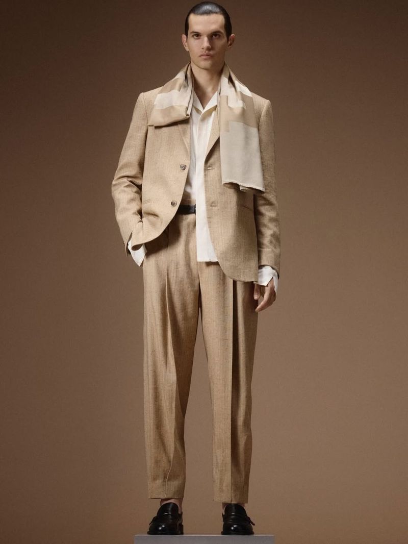 Model standing on a plinth wearing a tailored beige suit, cream shirt, draped scarf, and black loafers against a brown studio backdrop.