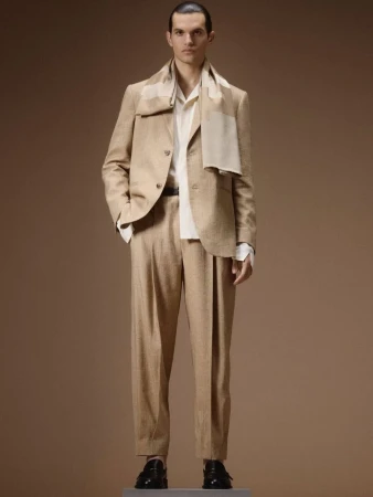 Model standing on a plinth wearing a tailored beige suit, cream shirt, draped scarf, and black loafers against a brown studio backdrop.