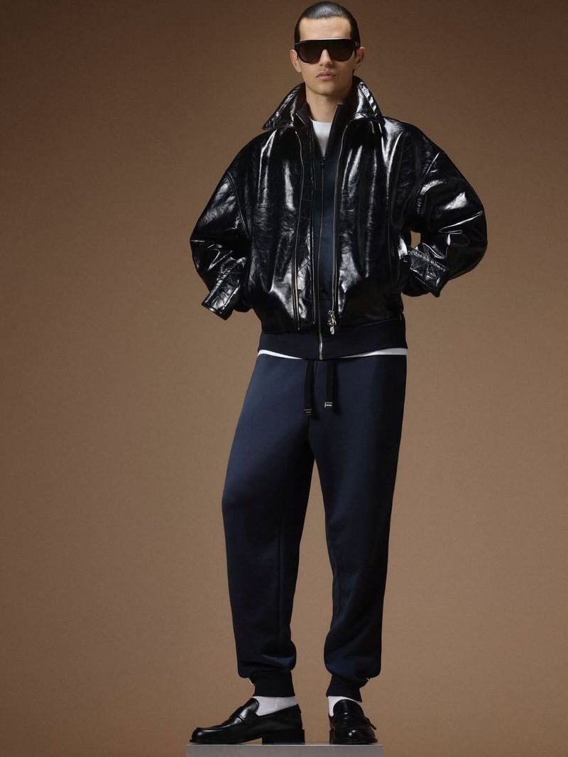 Male model wearing dark sunglasses, a glossy black bomber jacket, and navy cuffed track pants against a camel brown backdrop.