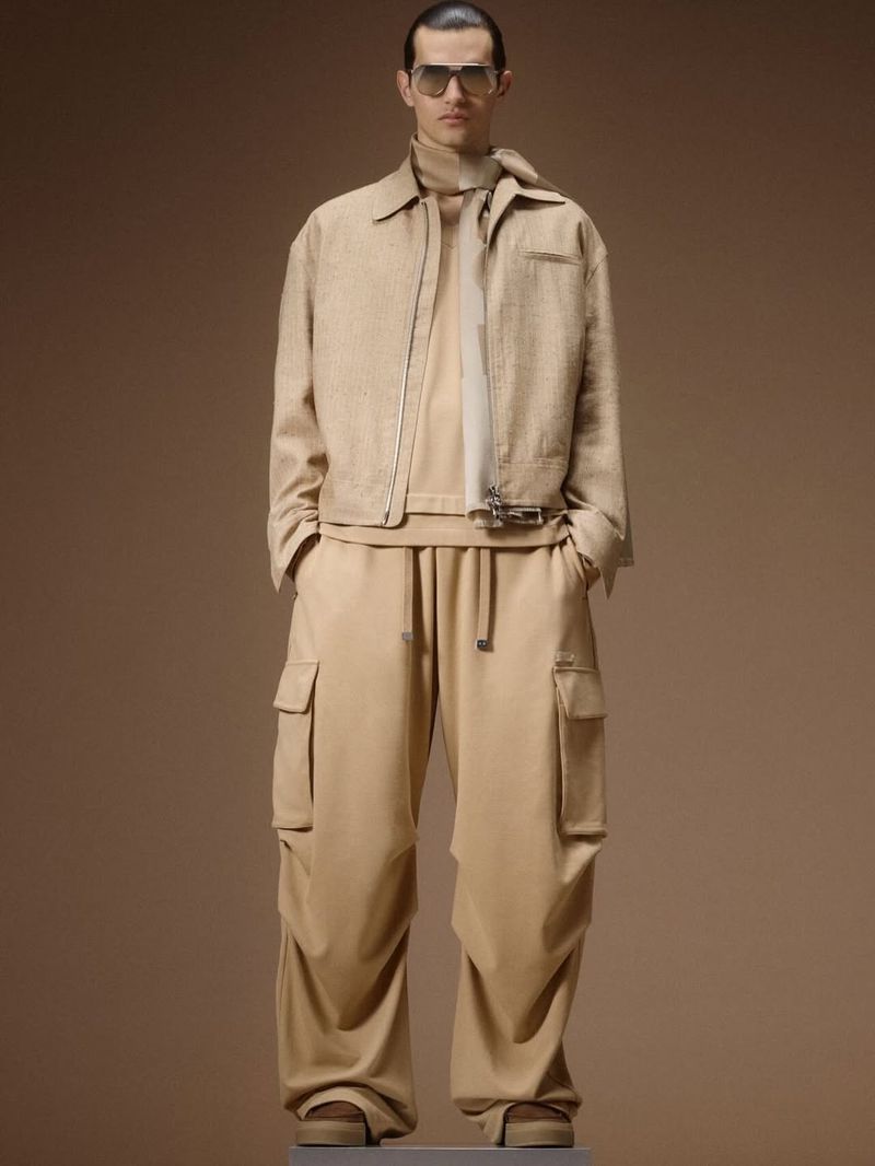 Male model wearing a monochromatic beige zip jacket, oversized cargo pants, neck scarf, and sunglasses against a warm tan backdrop.