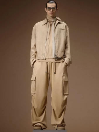 Male model wearing a monochromatic beige zip jacket, oversized cargo pants, neck scarf, and sunglasses against a warm tan backdrop.