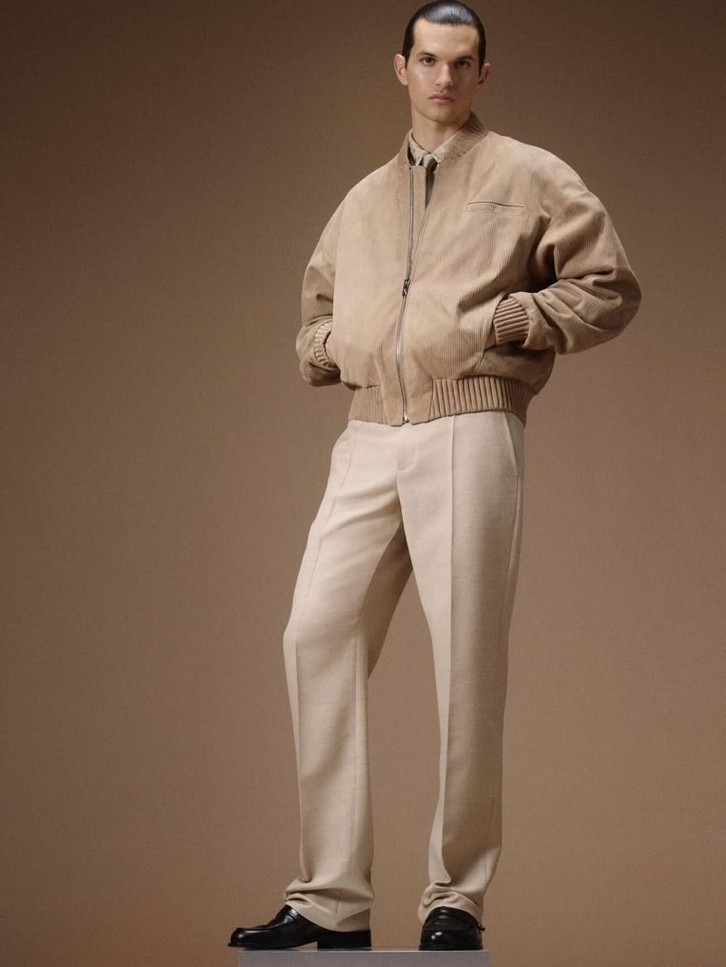 Model standing in a tan bomber jacket and light trousers against a neutral studio backdrop