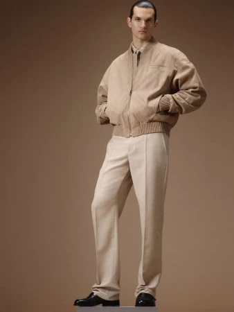 Model standing in a tan bomber jacket and light trousers against a neutral studio backdrop