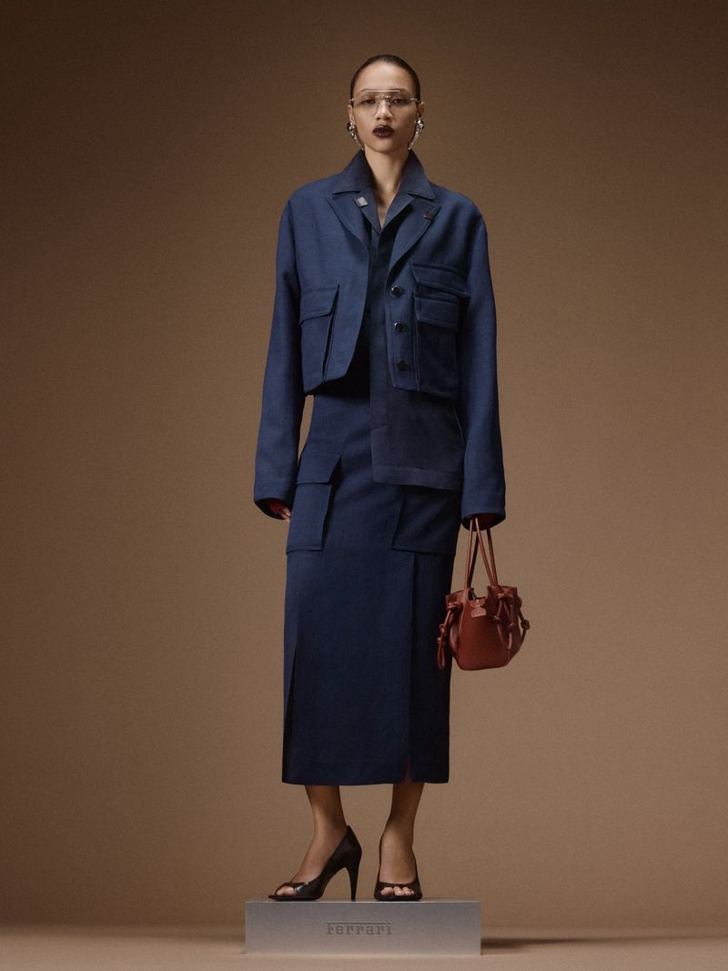 Model wearing a navy blue cropped utility jacket and matching skirt holding a small brown handbag against a beige studio backdrop.