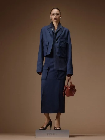 Model wearing a navy blue cropped utility jacket and matching skirt holding a small brown handbag against a beige studio backdrop.