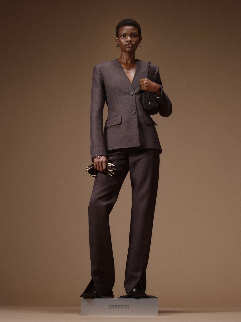 Model standing on a plinth wearing a structured dark brown suit holding a handbag against a seamless brown background.