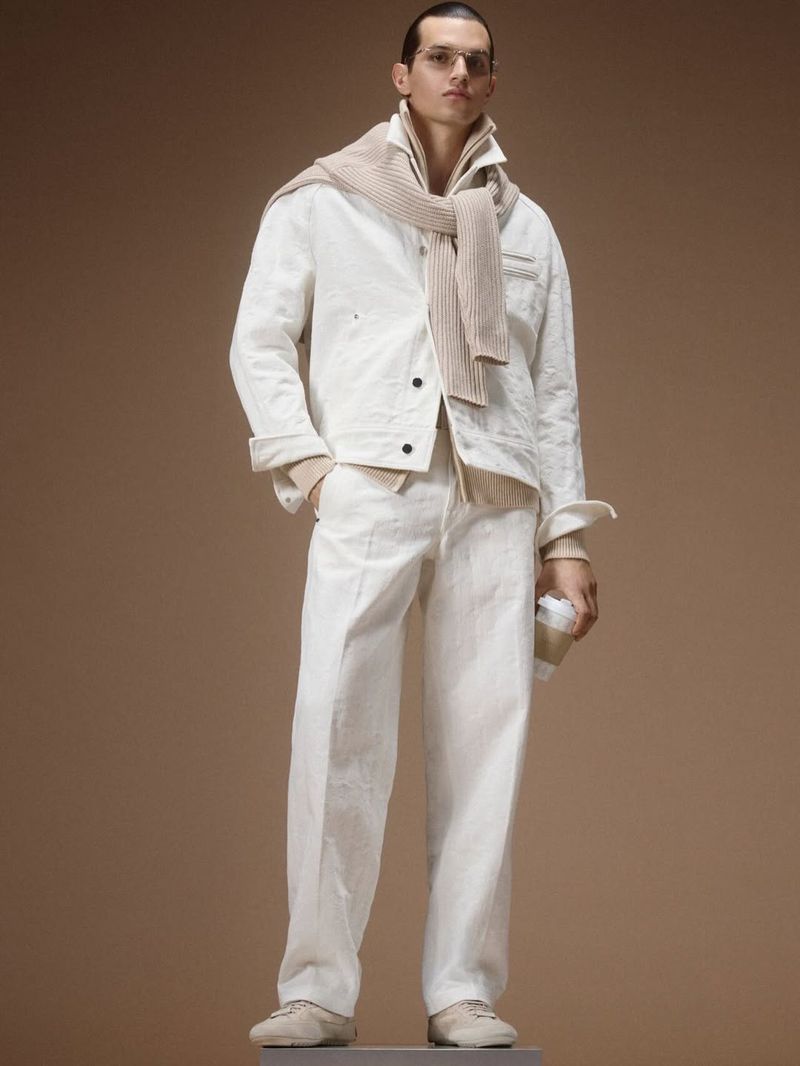 Male model posing in a white jacket and matching trousers with a beige knit sweater draped over shoulders against a tan backdrop.