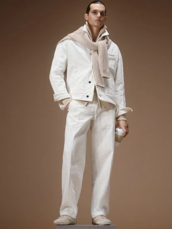 Male model posing in a white jacket and matching trousers with a beige knit sweater draped over shoulders against a tan backdrop.
