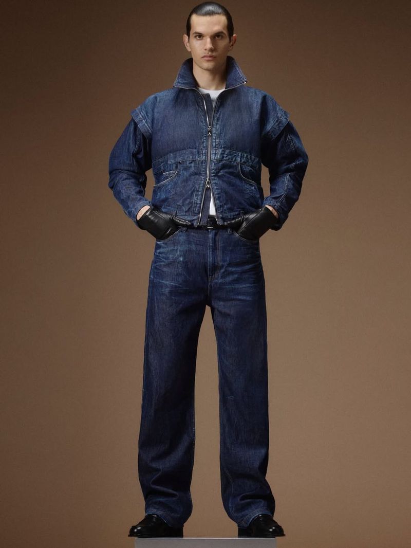 Model wearing a dark denim jacket, matching jeans, and black leather gloves against a brown seamless background.