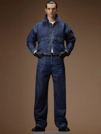 Model wearing a dark denim jacket, matching jeans, and black leather gloves against a brown seamless background.