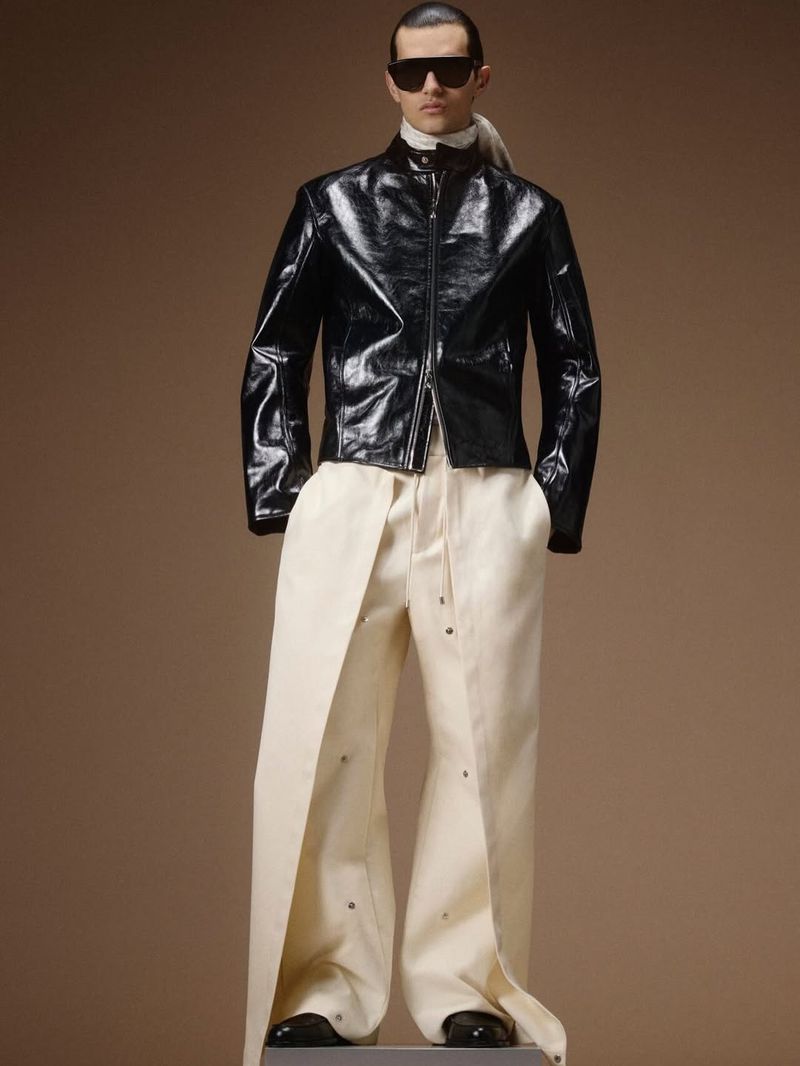 Model wearing a glossy black leather zip-up jacket, dark sunglasses, and wide-leg cream trousers against a warm beige background.