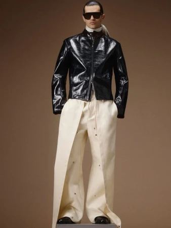 Model wearing a glossy black leather zip-up jacket, dark sunglasses, and wide-leg cream trousers against a warm beige background.