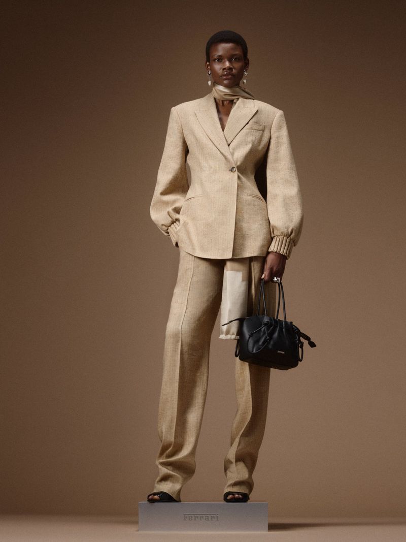 Model wearing a tailored beige pantsuit holding a black leather handbag on a branded plinth against a warm brown backdrop.