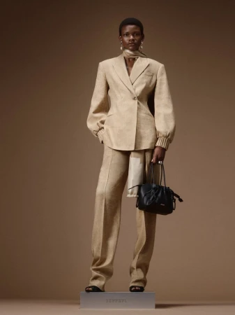 Model wearing a tailored beige pantsuit holding a black leather handbag on a branded plinth against a warm brown backdrop.
