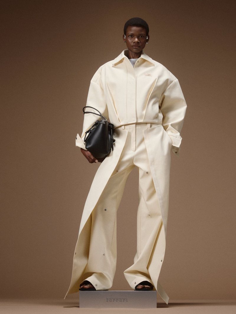 Model posing in an off-white structured jacket and draped trousers with a black handbag against a brown studio backdrop.