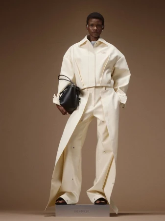 Model posing in an off-white structured jacket and draped trousers with a black handbag against a brown studio backdrop.