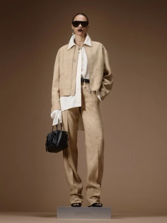 Model wearing a beige jacket, matching wide-leg trousers, white collared shirt, sunglasses, and holding a small black handbag.