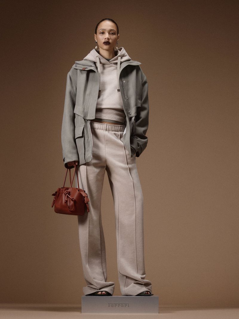Model standing on a plinth in a grey oversized jacket, matching hooded sweatshirt, wide-leg sweatpants, and holding a brown bag.