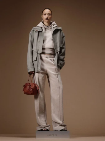 Model standing on a plinth in a grey oversized jacket, matching hooded sweatshirt, wide-leg sweatpants, and holding a brown bag.