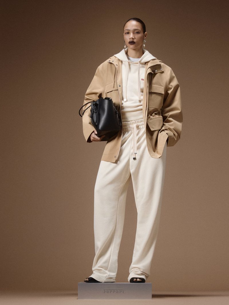 Model posing in a tan utility jacket and cream tracksuit on a low pedestal with a black bag against a beige studio backdrop.