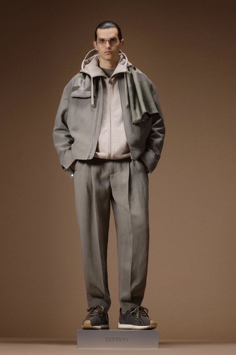 Male model in a taupe layered zip jacket and pleated trousers standing on a branded plinth against a warm brown backdrop.