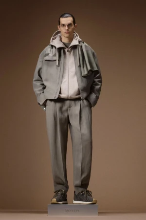 Male model in a taupe layered zip jacket and pleated trousers standing on a branded plinth against a warm brown backdrop.