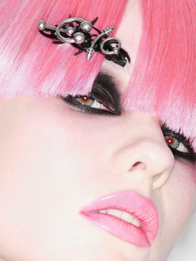 Close-up beauty portrait of model Lola Varley with straight pink bangs, bold black eye makeup, and a silver and black hair clip.