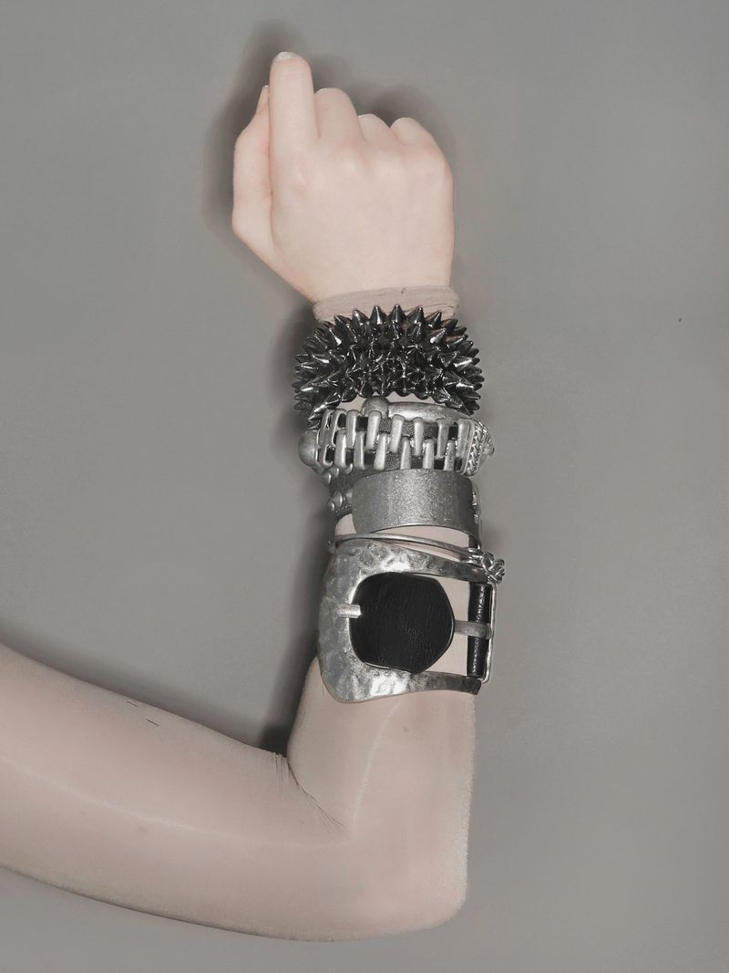 Arm wearing a stack of chunky metallic bracelets and cuffs against a neutral grey background for Minga London.