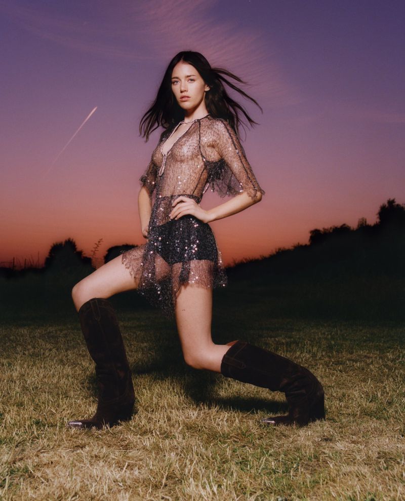 A woman with long dark hair poses outdoors in a sheer black dress and high boots against a purple and orange sky backdrop.