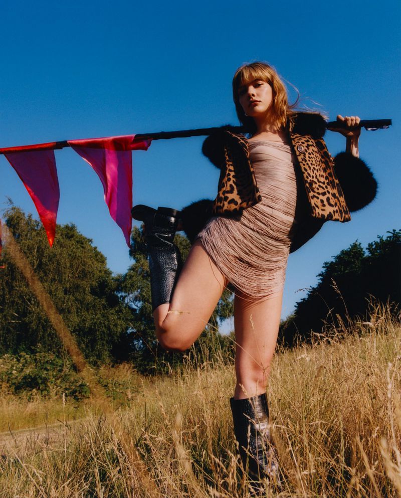 Adult woman in a dress with fringe and a spotted jacket standing in a field of dry grass, holding a pole with pink flags. 