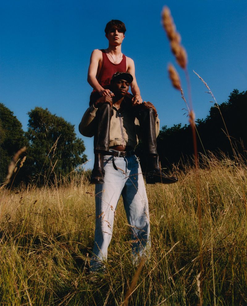 Two men pose in a grassy field on a sunny day. One man is giving the other a piggyback ride in the meadow.
