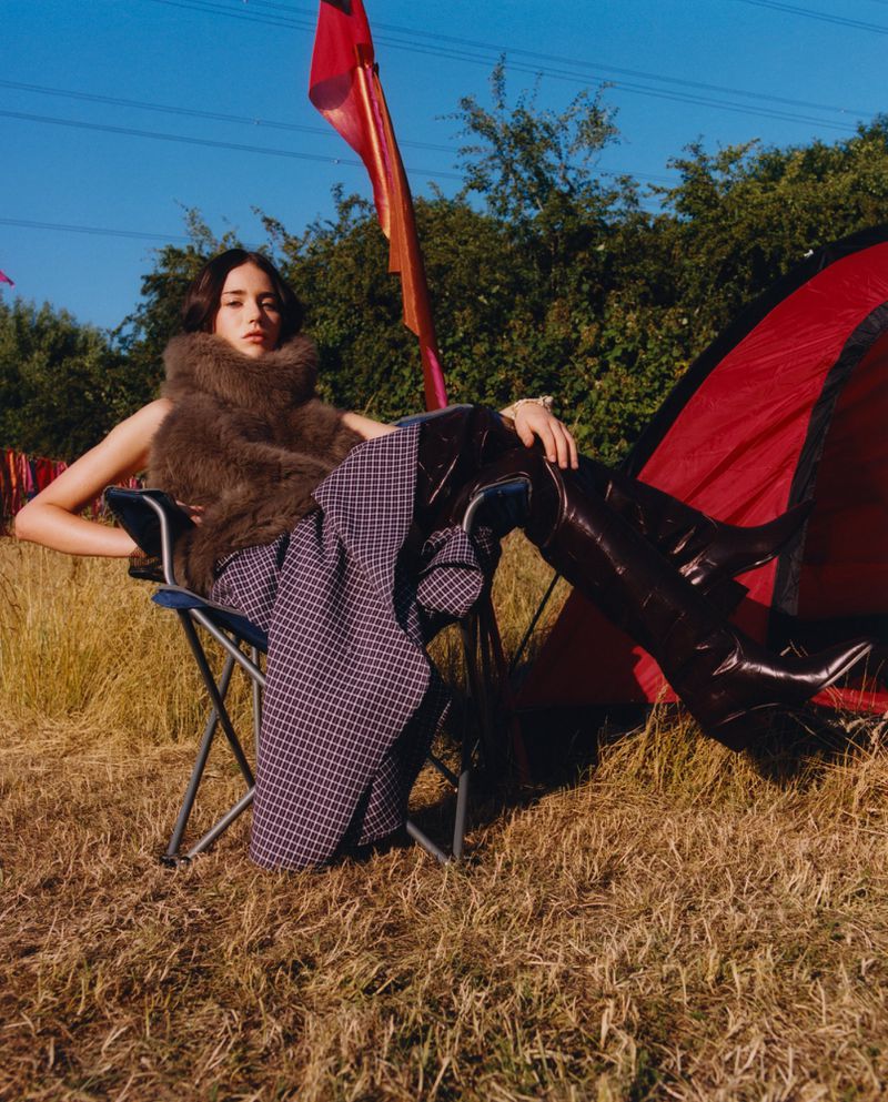 A model is sitting in a chair on a grassy field, wearing a fur vest and thigh-high boots, with a tent and trees in the background.