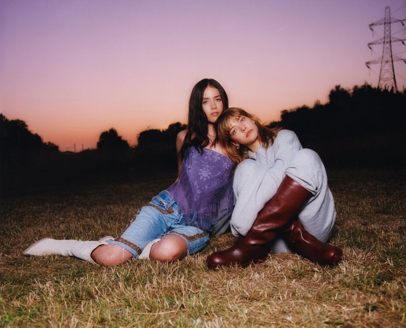 Two women pose in a grassy field at dusk wearing casual clothing and boots, with an electrical tower visible in the distance.