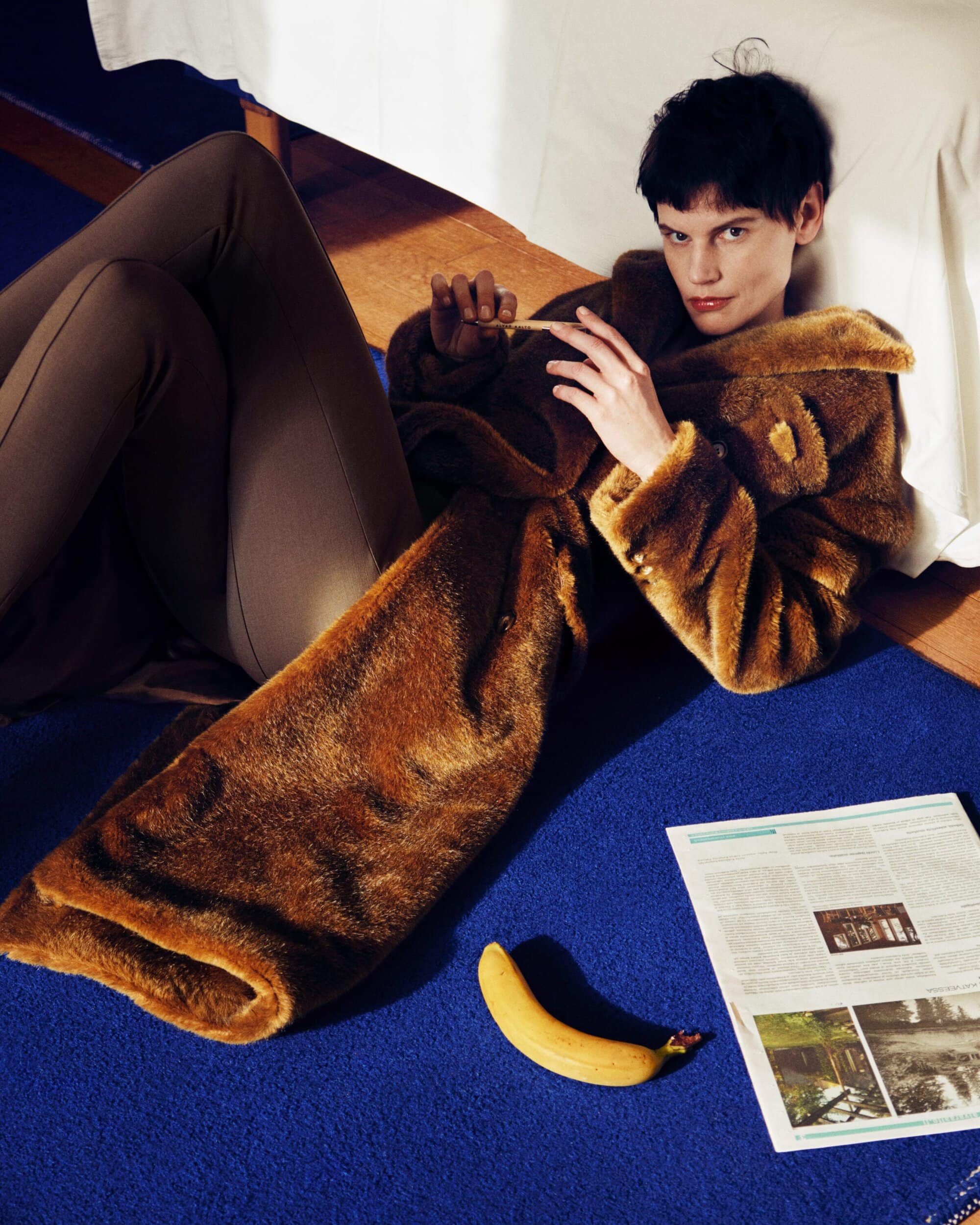 Model Saskia de Brauw reclining on a deep blue carpet in a brown faux fur coat beside a banana and an open newspaper.