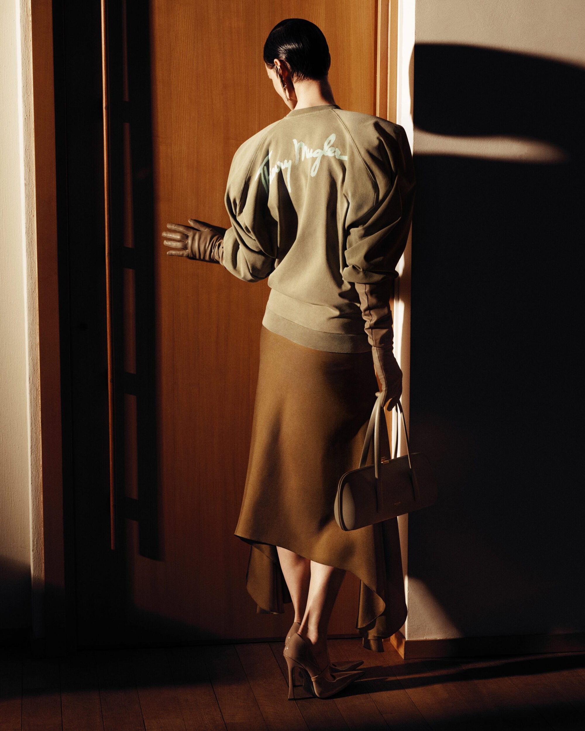 Model Saskia de Brauw from behind wearing an embroidered taupe sweatshirt and asymmetric skirt by a wooden door in hard light.