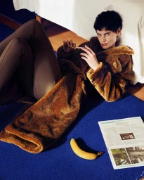 Model Saskia de Brauw reclining on a deep blue carpet in a brown faux fur coat beside a banana and an open newspaper.