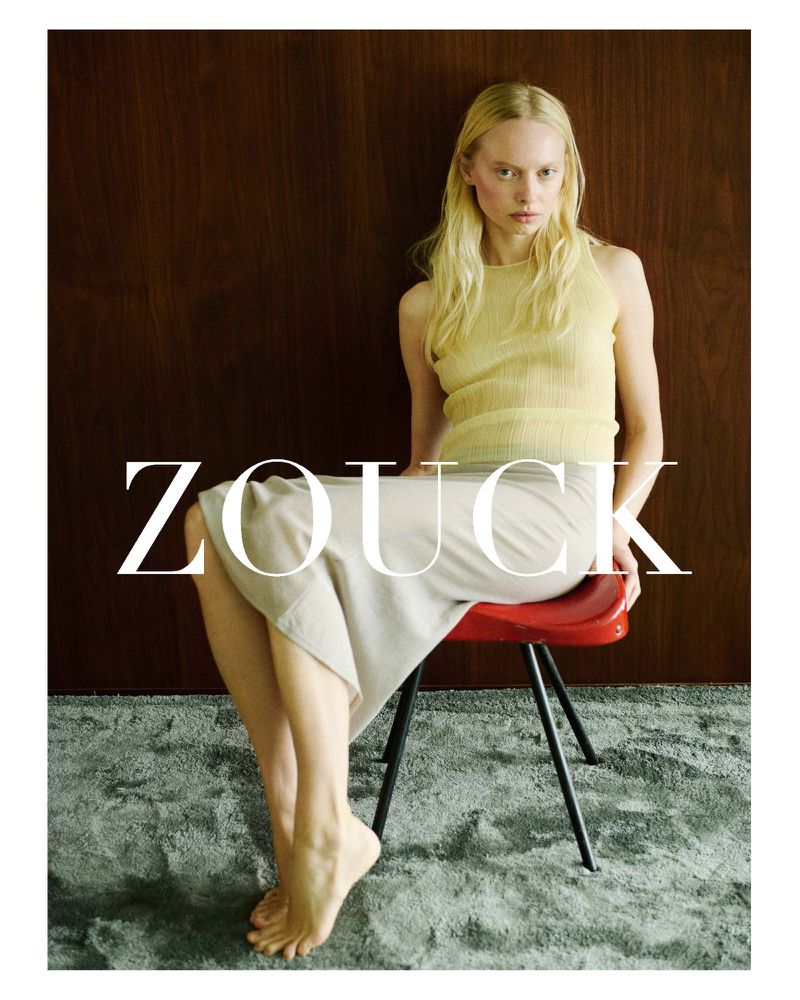 Model India Tuersley seated barefoot on a red chair wearing a sheer yellow top and off-white midi skirt against a dark wood wall.
