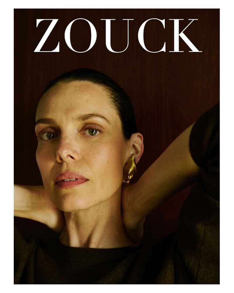 Model India Tuersley wearing a sculptural gold earring and a dark sweater with hands behind neck, beneath white text reading ZOUCK.