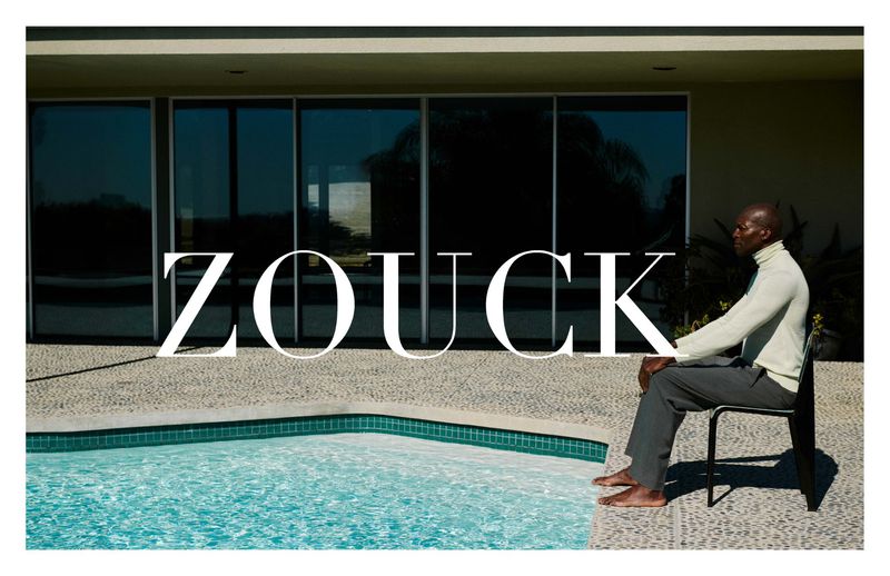 A man in an off-white turtleneck and grey trousers sits barefoot on a chair beside a turquoise pool in front of a modern house.