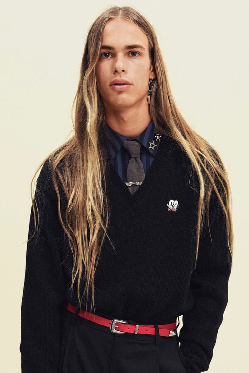 Model with long blonde hair wearing a black V-neck sweater over a striped blue shirt against a neutral background