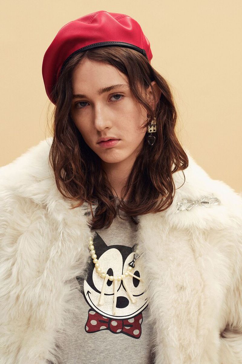 Model wearing a bright red beret and plush white faux fur coat over a graphic top against a warm studio backdrop.