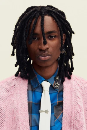 Portrait of a Black man with dreadlocks wearing a pink knit cardigan, blue plaid shirt, and cream tie against a plain backdrop.
