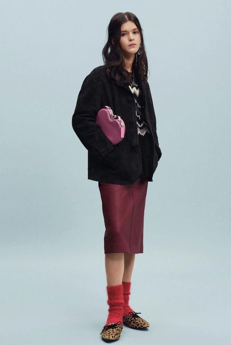 Model wearing a black jacket, burgundy midi skirt, leopard flats, and holding a pink heart bag against a light blue backdrop.